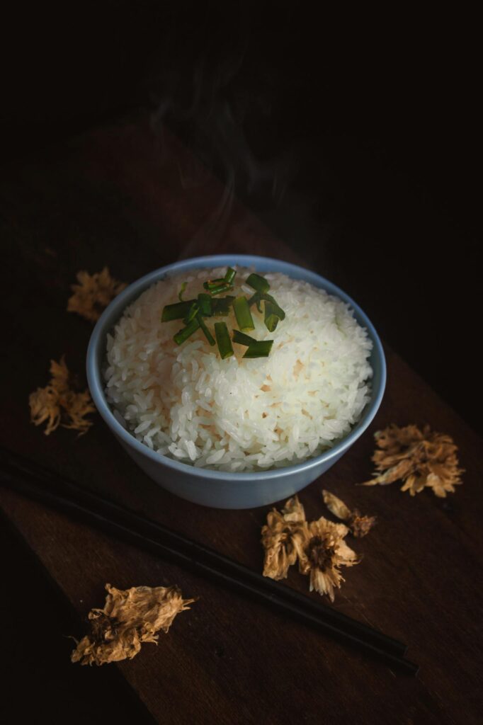 A steaming bowl of rice garnished with green onions, surrounded by dried flowers, captured in a rustic style.