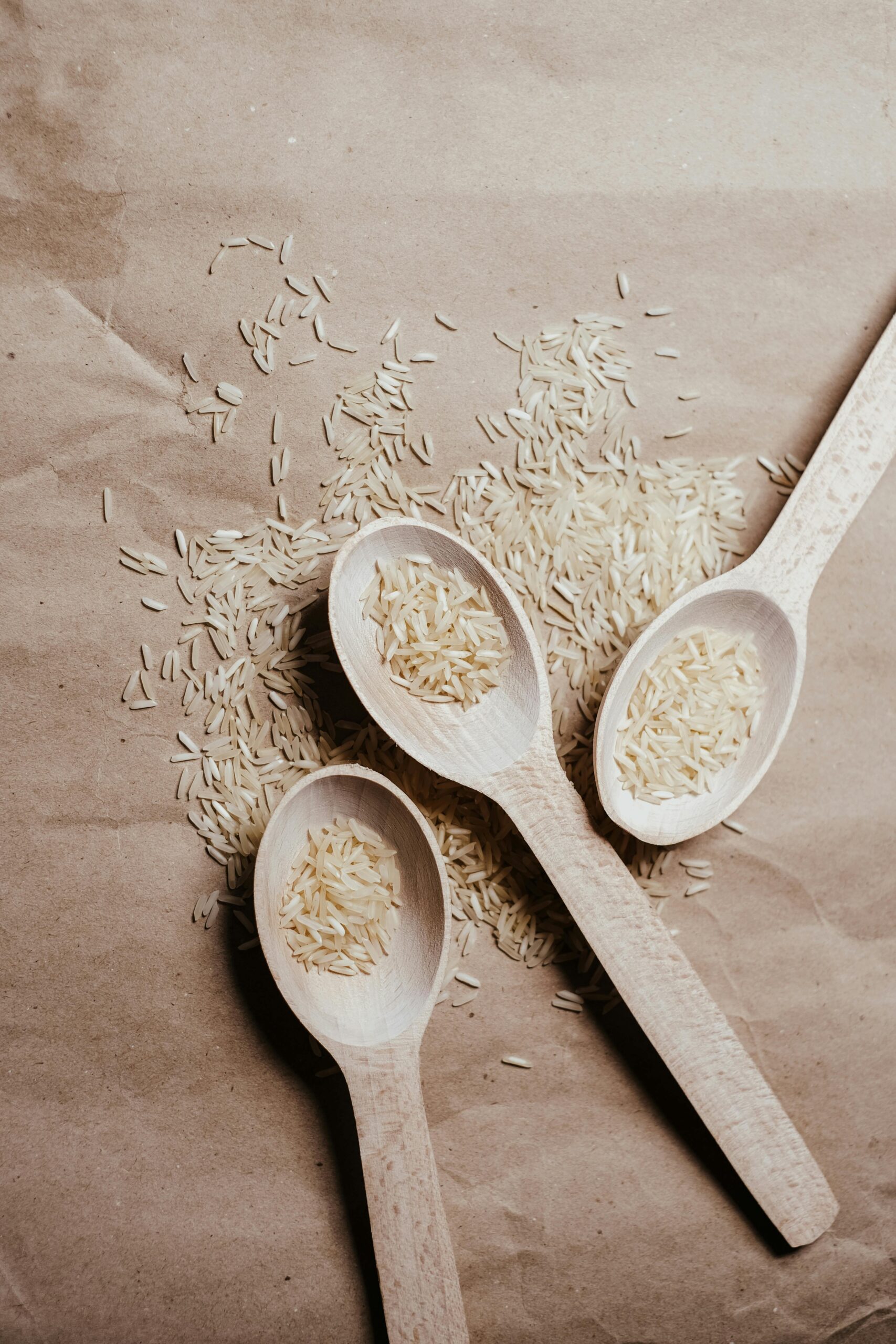 Flat lay image of uncooked rice on wooden spoons on a brown paper surface, perfect for food blogs.